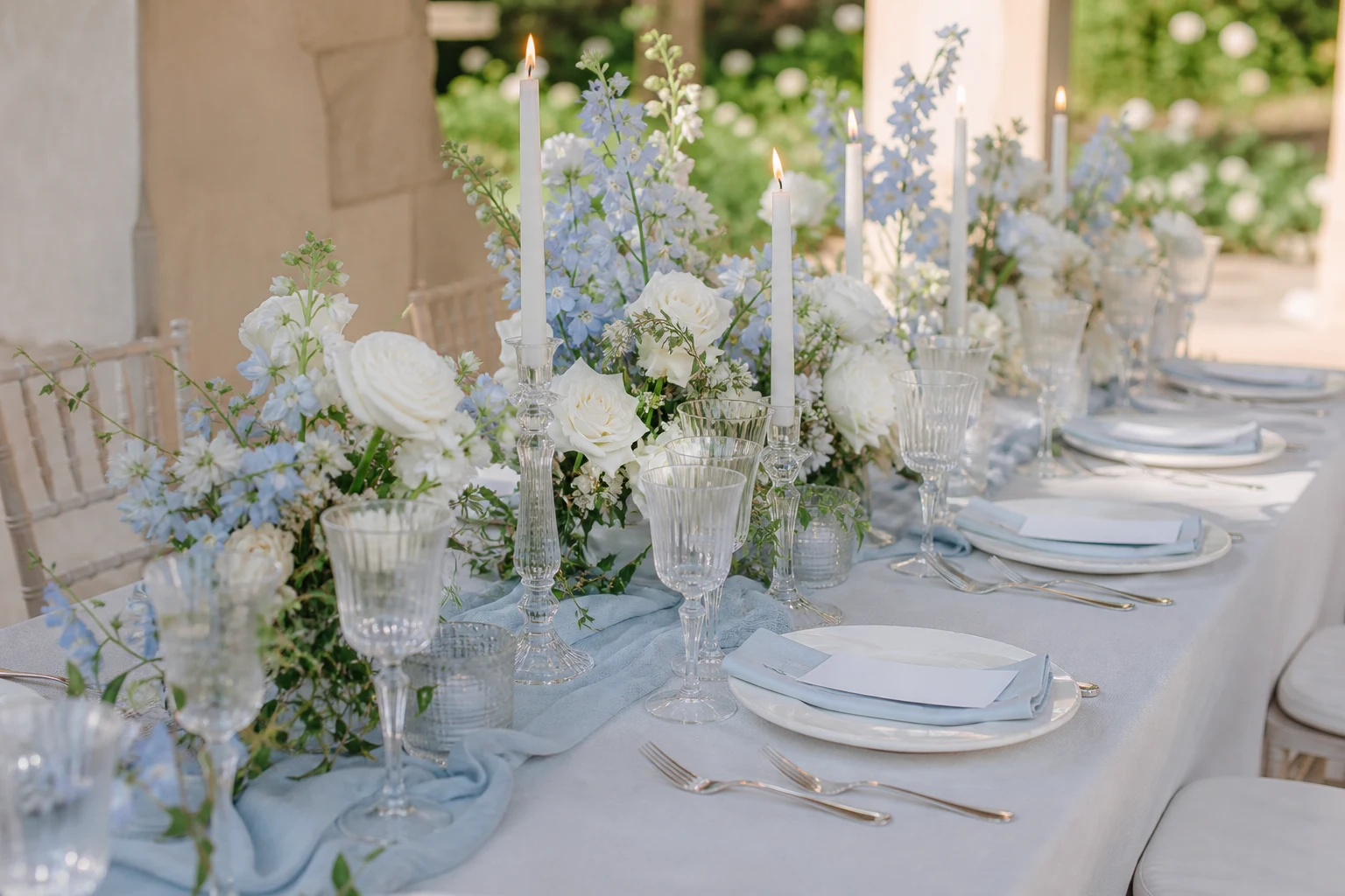 Elegant outdoor wedding table with powder-blue linens, white flowers, glassware, and taper candles