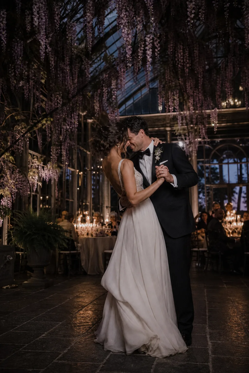 Newlyweds dancing beneath hanging wisteria in a candlelit glass conservatory