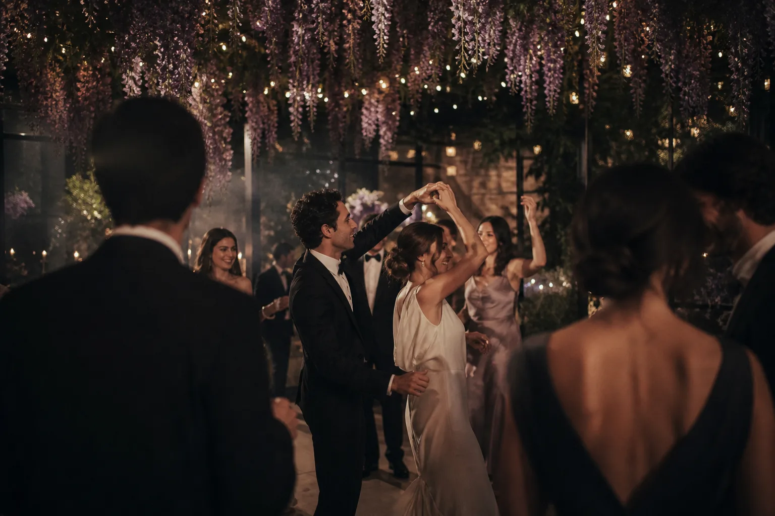 Newlyweds and guests dancing beneath wisteria and warm lights at an evening reception